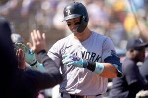 Aaron Judge, standing in the dugout, smiles while watching his teammates warm up before the game. The Yankees slugger remains focused as he enters the final week of the season, leading the home run race against Shohei Ohtani.